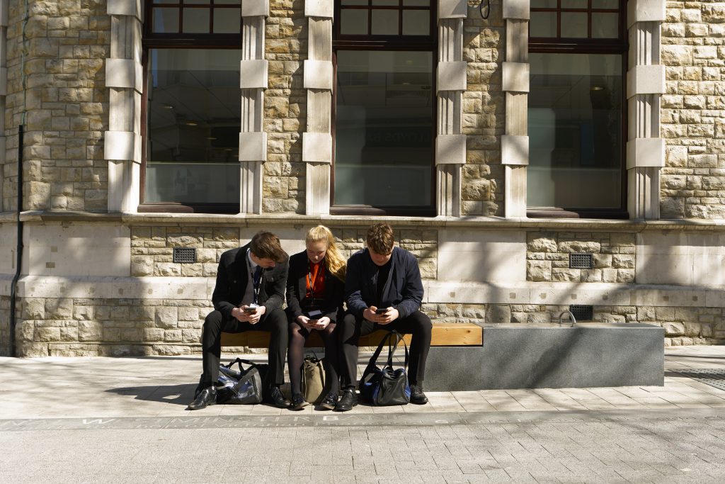 Secondary school aged teenagers sitting on a bench on a sunny day with a stone building in the background